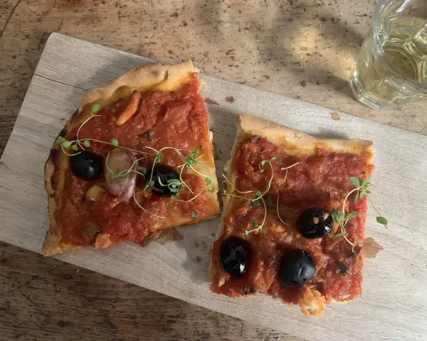 Zwei Scheiben Focaccia mit Tomaten, Knoblauch und schwarzen **Oliven Ligurien** garniert mit frischen Kräutern, daneben ein Glas Weißwein.