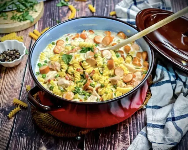 Herzhafte Nudelpfanne mit Würstchen und Erbsen, perfekt für schnelle Abendbrot Rezepte. Ein Holzlöffel liegt bereit.