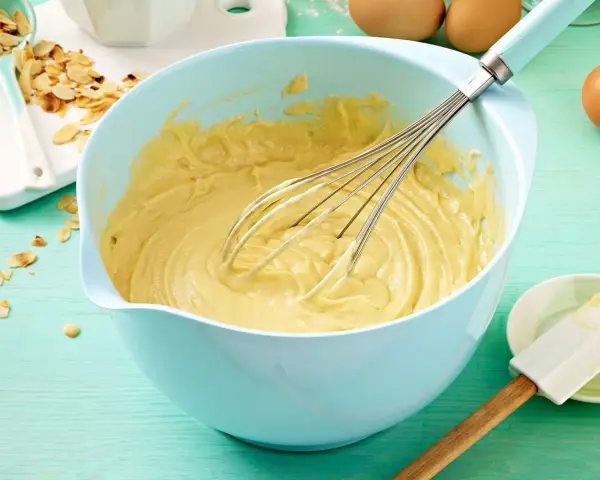 Ein hellblauer Schüssel mit Kuchenteig und Schneebesen. Daneben Mandelsplitter und Eier. Ein tolles Kuchenteig Rezept!