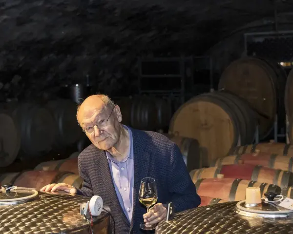 Ein Mann mit Brille hält ein Glas Wein in einem Weinkeller. Die Fässer im Hintergrund und der Wein lassen an Wein und Musik denken.