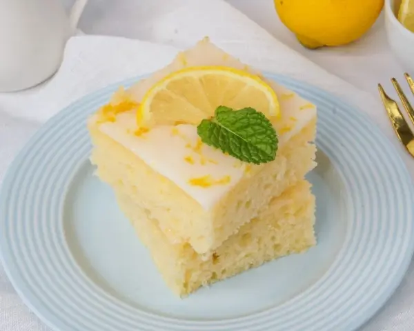 Zwei Stücke griechischer Zitronenkuchen mit Joghurt, garniert mit Zitronenscheibe und Minze, auf einem hellblauen Teller.