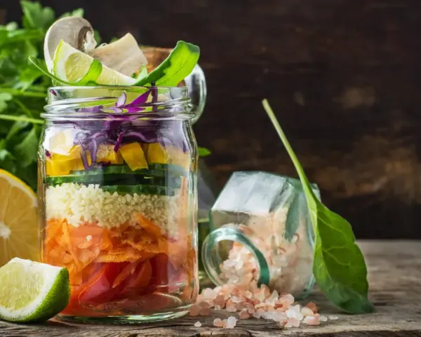 Bunter Salat im Glas mit Couscous, Karotten, Gurken, Paprika und Rotkohl. Daneben liegen Zitronen und Kräuter.
