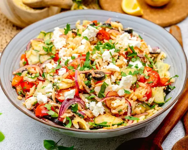 Bunter Kritharaki Salat mit Feta, Tomaten, Gurken, roten Zwiebeln und Oliven, garniert mit Petersilie. Ein köstlicher mediterraner Genuss.