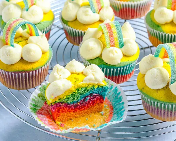 Bunte regenbogen muffins mit weißem Frosting und Gummibärchen-Regenbogen-Deko auf einem Gitter.