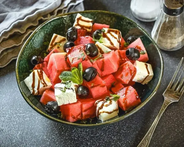 Erfrischender Melone Feta Salat mit schwarzen Oliven und Minze, beträufelt mit Balsamico-Glasur.