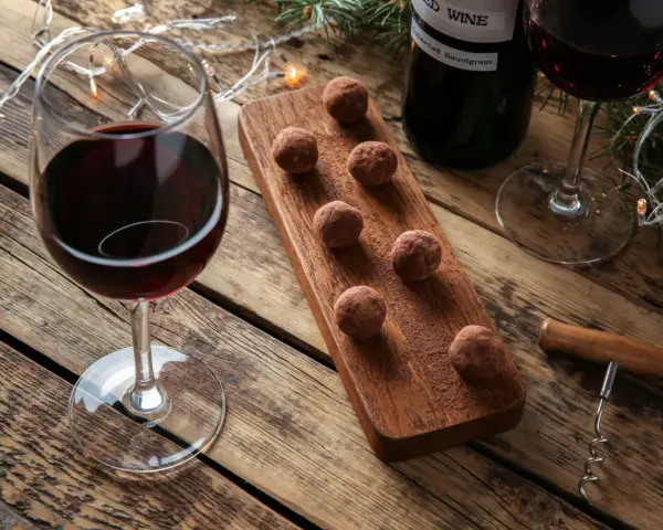 Ein Glas Rotwein und Schokolade-Trüffel auf einem Holzbrett, daneben eine Flasche Rotwein und Lichterketten.