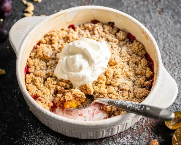 Ein warmer Zwetschgen Crumble mit Haferflocken, gekrönt von einem Klecks Sahne. Ein Löffel hebt die süße Frucht hervor.