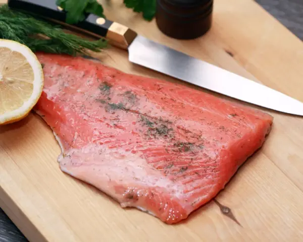 Frisches Lachsfilet mit Dill und Zitrone auf einem Holzbrett, bereit zum Kochen mit Gin. Ein scharfes Messer liegt daneben.
