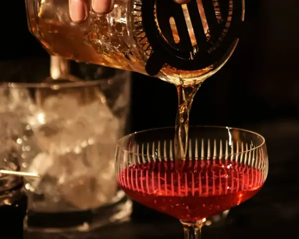 Ein **cocktail klassiker mit whisky und wermut** wird zubereitet. Bernstein-farbene Flüssigkeit fließt aus einem Shaker in ein Glas mit rotem Inhalt.