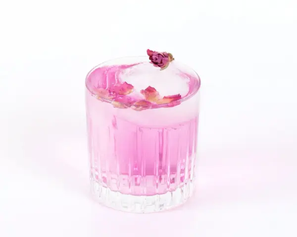 Ein erfrischender Gin Mix in zartem Rosa, garniert mit Rosenblättern und einer kleinen Knospe auf einem Eiswürfel.