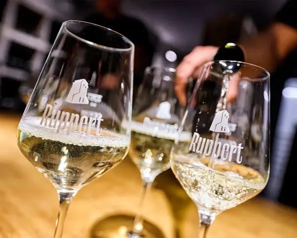 Drei Gläser mit schäumendem Wein, der von einer Flasche in das vorderste Glas gegossen wird. Der Wein aus mehreren Rebsorten ist von der Marke Ruppert.