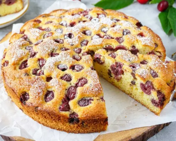 Ein saftiger, sommerlicher Kuchen mit Kirschen, bestäubt mit Puderzucker. Ein Stück wird gerade herausgeschnitten.