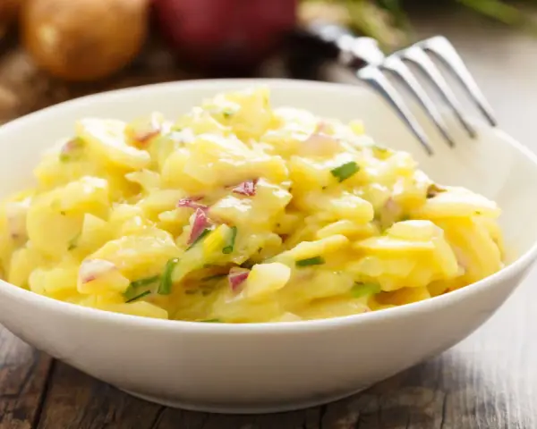 Ein cremiger Kartoffelsalat mit roten Zwiebeln und Schnittlauch in einer weißen Schüssel, bereit zum Genießen. Perfekt für ein Picknick oder als Beilage.
