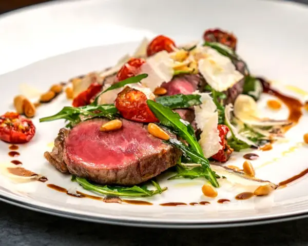 Saftiges Tagliata di Manzo mit Rucola, Kirschtomaten, Pinienkernen und Parmesan auf einem weißen Teller.