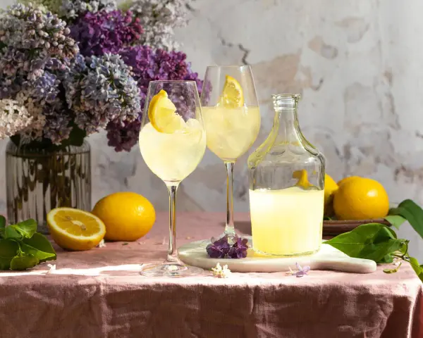 Zwei Gläser mit Eis und Zitronenscheiben, eine Flasche Limoncello und frische Zitronen auf einem rosafarbenen Tischtuch. Ein Hauch von Frühling mit Flieder.