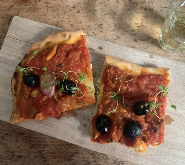 Zwei Scheiben Focaccia mit Tomaten, Knoblauch und schwarzen **Oliven Ligurien** garniert mit frischen Kräutern, daneben ein Glas Weißwein.