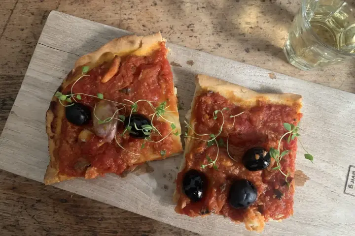 Zwei Scheiben Focaccia mit Tomaten, Knoblauch und schwarzen **Oliven Ligurien** garniert mit frischen Kräutern, daneben ein Glas Weißwein.