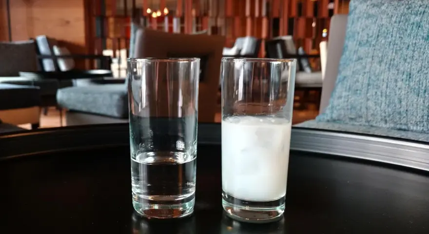 Zwei Gläser auf einem Tisch: eines mit klarem Wasser, das andere mit milchigem Ouzo. Ein Vergleich zwischen Raki und Ouzo.