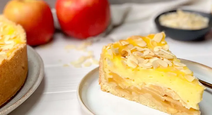 Ein saftiges Stück Apfel Schmand Torte mit Mandelsplittern, daneben Äpfel und eine Schale mit Streuseln.