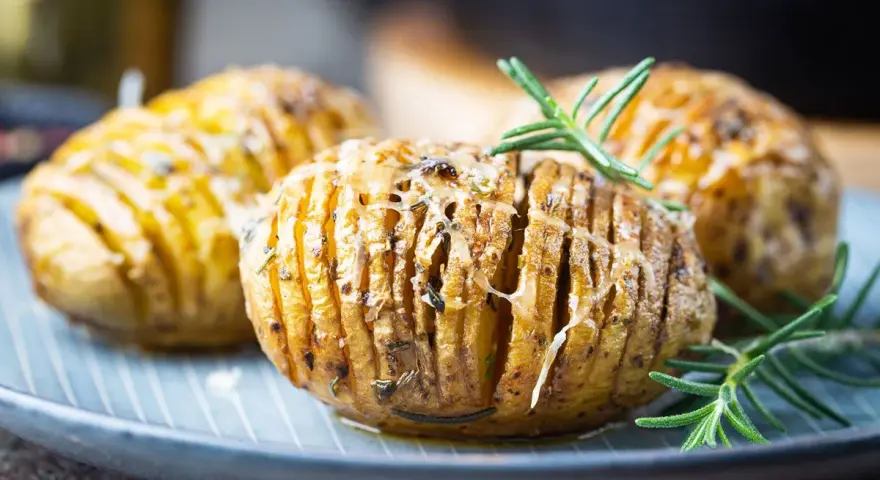 Drei goldbraune Hasselback Kartoffeln mit geschmolzenem Käse und Rosmarin auf einem blauen Teller.