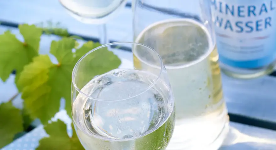 Erfrischendes Weinschorle-Rezept: Weißwein und Mineralwasser in Gläsern, daneben eine Karaffe und eine Flasche Mineralwasser. Grüne Weinblätter im Hintergrund.