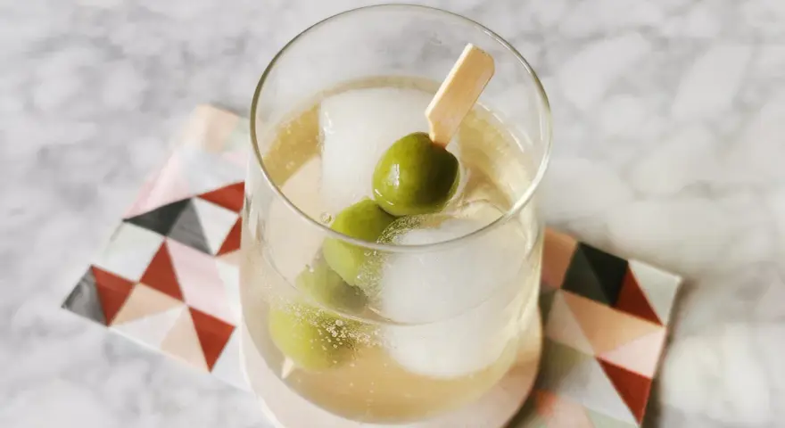 Erfrischender Italicus Spritz mit Eiswürfeln und Oliven auf einem gemusterten Untersetzer.