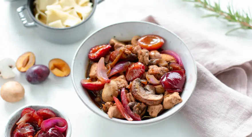 Herzhafte Rezepte mit Pflaumen: Ein Eintopf mit Fleisch, Pilzen und roten Zwiebeln, garniert mit frischen Pflaumen. Daneben eine Schüssel mit Nudeln.