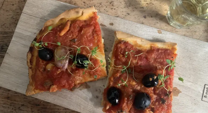 Zwei Scheiben Focaccia mit Tomaten, Knoblauch und schwarzen **Oliven Ligurien** garniert mit frischen Kräutern, daneben ein Glas Weißwein.