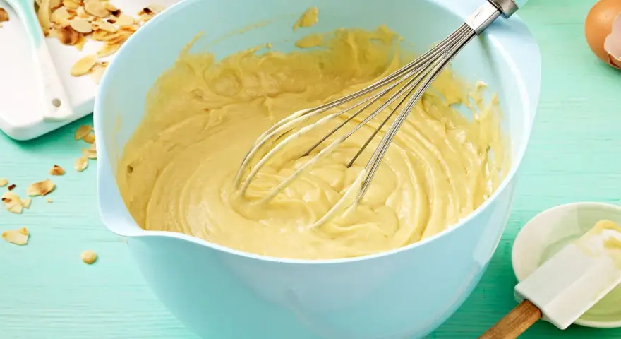 Ein hellblauer Schüssel mit Kuchenteig und Schneebesen. Daneben Mandelsplitter und Eier. Ein tolles Kuchenteig Rezept!