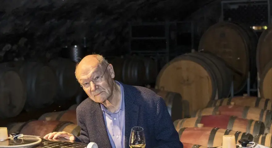 Ein Mann mit Brille hält ein Glas Wein in einem Weinkeller. Die Fässer im Hintergrund und der Wein lassen an Wein und Musik denken.