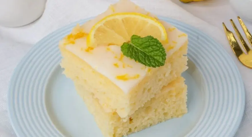 Zwei Stücke griechischer Zitronenkuchen mit Joghurt, garniert mit Zitronenscheibe und Minze, auf einem hellblauen Teller.