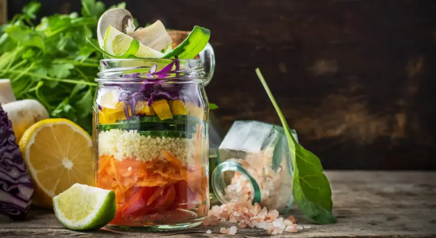 Bunter Salat im Glas mit Couscous, Karotten, Gurken, Paprika und Rotkohl. Daneben liegen Zitronen und Kräuter.