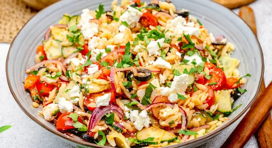 Bunter Kritharaki Salat mit Feta, Tomaten, Gurken, roten Zwiebeln und Oliven, garniert mit Petersilie. Ein köstlicher mediterraner Genuss.