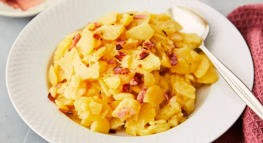 Ein Teller mit Kartoffelsalat mit Speck, garniert mit knusprigen Speckwürfeln. Daneben liegt ein Löffel.