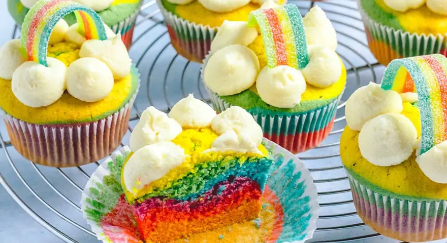 Bunte regenbogen muffins mit weißem Frosting und Gummibärchen-Regenbogen-Deko auf einem Gitter.