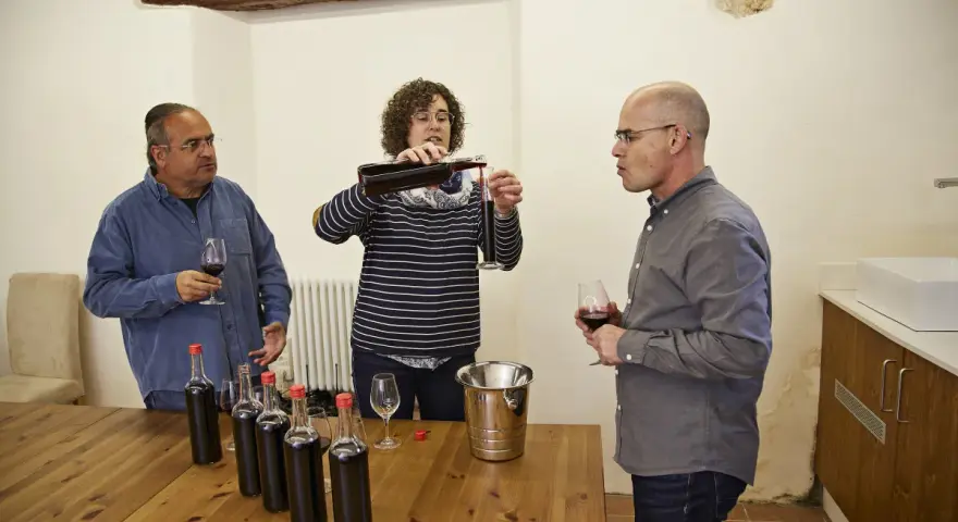 Weinprobe: Eine Frau schenkt Wein ein, während zwei Männer zuschauen. Auf dem Tisch stehen Flaschen für die Assemblage.