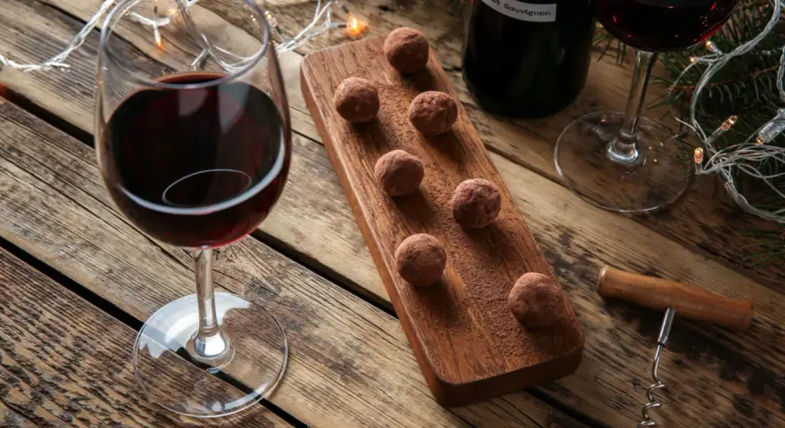 Ein Glas Rotwein und Schokolade-Trüffel auf einem Holzbrett, daneben eine Flasche Rotwein und Lichterketten.