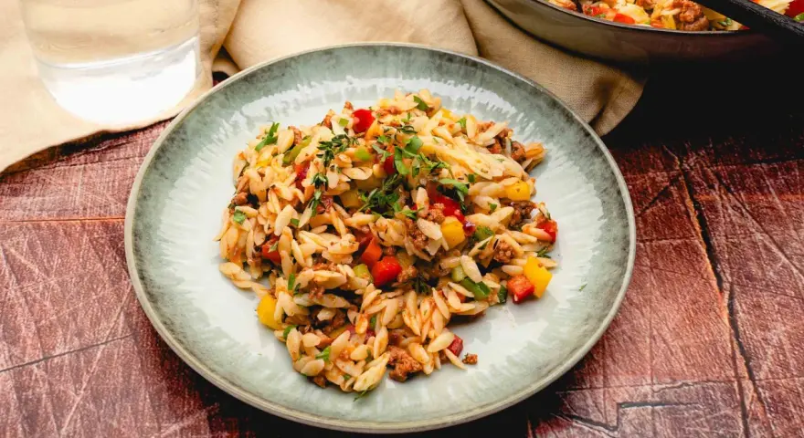 Ein Teller mit kritharaki salat mit hackfleisch, garniert mit frischen Kräutern. Daneben ein Glas Wasser und eine Pfanne mit dem Gericht.