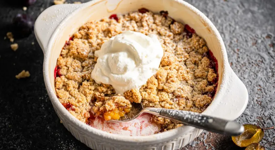 Ein warmer Zwetschgen Crumble mit Haferflocken, gekrönt von einem Klecks Sahne. Ein Löffel hebt die süße Frucht hervor.