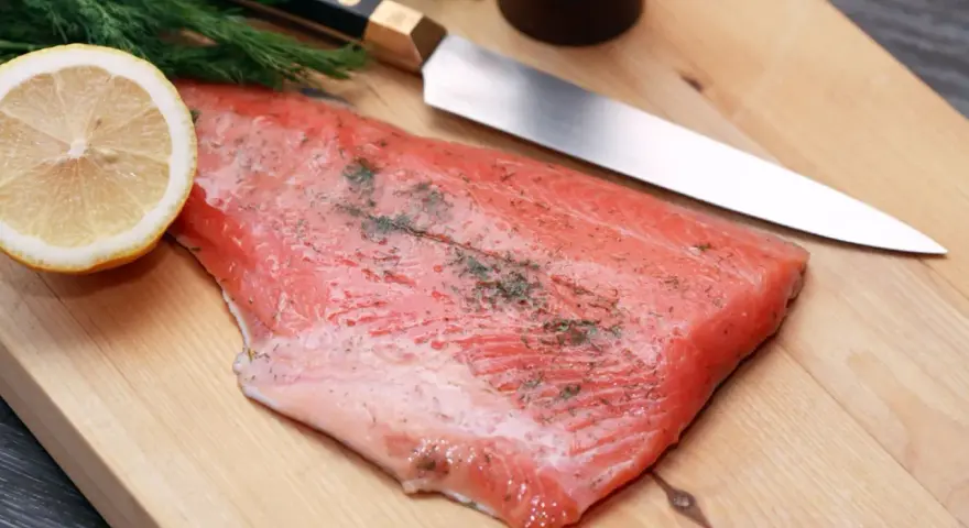Frisches Lachsfilet mit Dill und Zitrone auf einem Holzbrett, bereit zum Kochen mit Gin. Ein scharfes Messer liegt daneben.