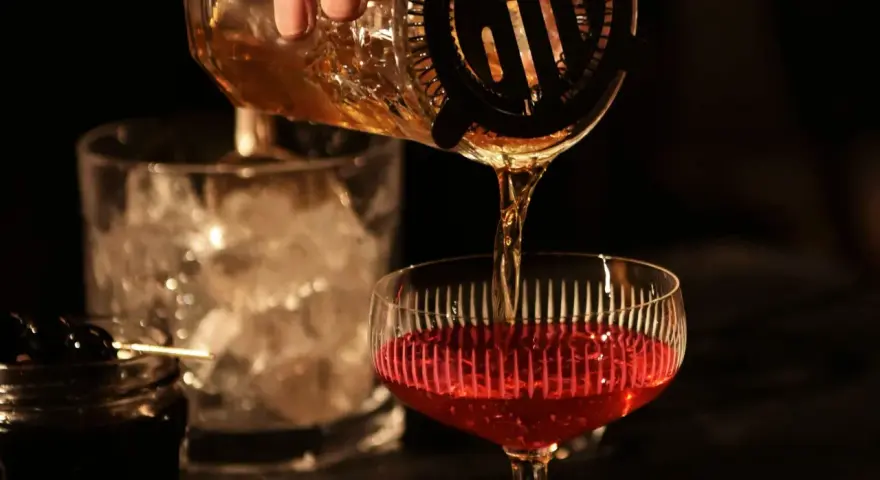 Ein **cocktail klassiker mit whisky und wermut** wird zubereitet. Bernstein-farbene Flüssigkeit fließt aus einem Shaker in ein Glas mit rotem Inhalt.