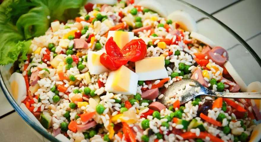 Reissalat mit Erbsen, Karotten, Ei und Wurst – ein gesunder Salat zum Grillen, garniert mit Tomaten und Salatblättern.