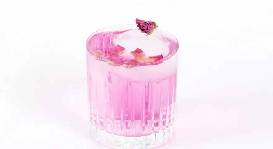 Ein erfrischender Gin Mix in zartem Rosa, garniert mit Rosenblättern und einer kleinen Knospe auf einem Eiswürfel.