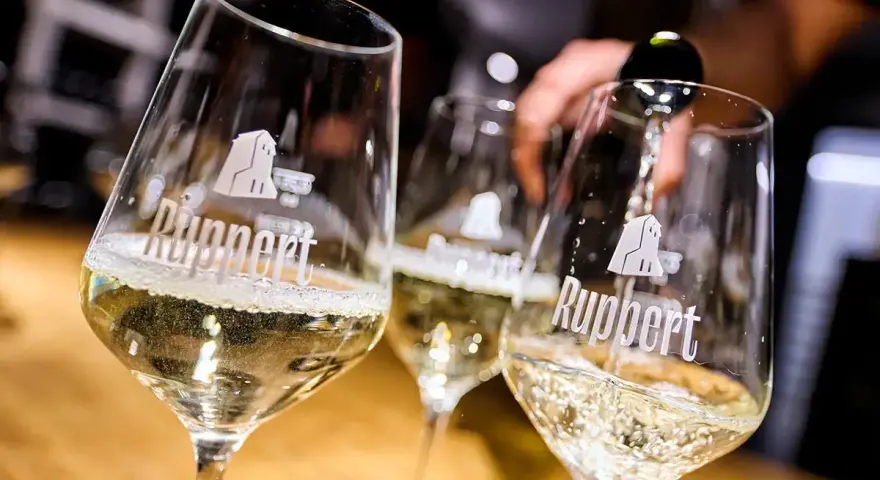 Drei Gläser mit schäumendem Wein, der von einer Flasche in das vorderste Glas gegossen wird. Der Wein aus mehreren Rebsorten ist von der Marke Ruppert.