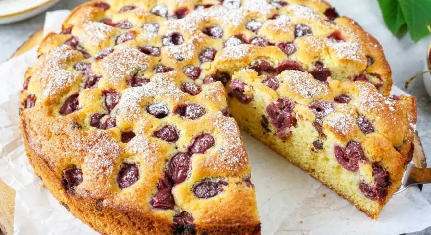 Ein saftiger, sommerlicher Kuchen mit Kirschen, bestäubt mit Puderzucker. Ein Stück wird gerade herausgeschnitten.