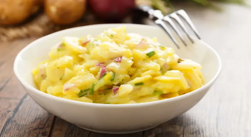 Ein cremiger Kartoffelsalat mit roten Zwiebeln und Schnittlauch in einer weißen Schüssel, bereit zum Genießen. Perfekt für ein Picknick oder als Beilage.