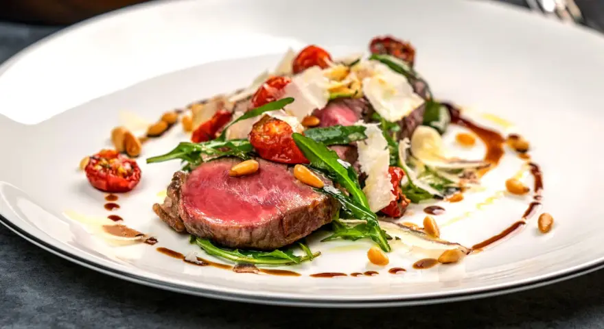 Saftiges Tagliata di Manzo mit Rucola, Kirschtomaten, Pinienkernen und Parmesan auf einem weißen Teller.