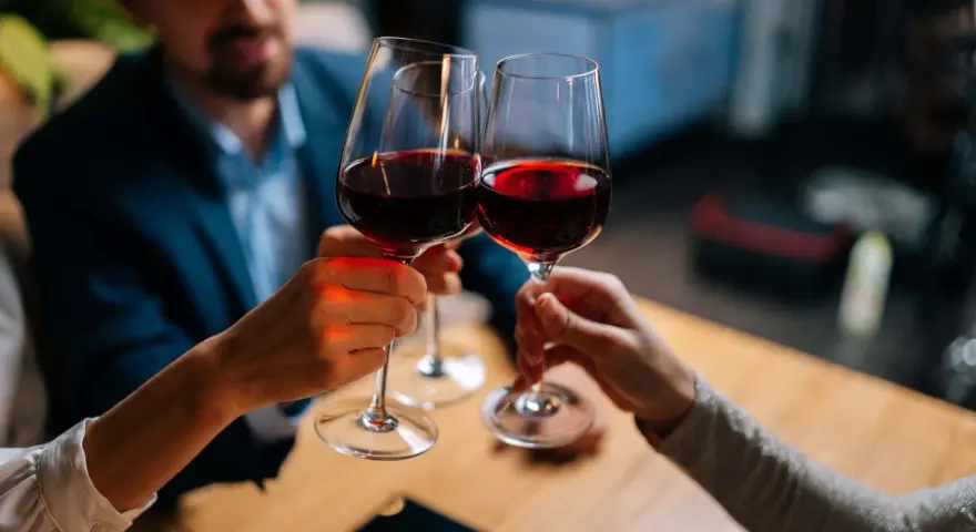 Drei Personen stoßen mit Rotwein an. Sie halten das weinglas richtig, am Stiel, um die Temperatur des Weins zu bewahren.