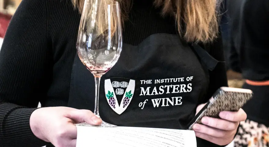 Eine Person mit einer Schürze des "Institute of Masters of Wine" hält ein Weinglas und ein Smartphone.