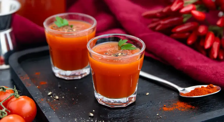Zwei Gläser würziger mexikanischer Tomatensaft, garniert mit Petersilie, neben frischen Tomaten und Chilis.